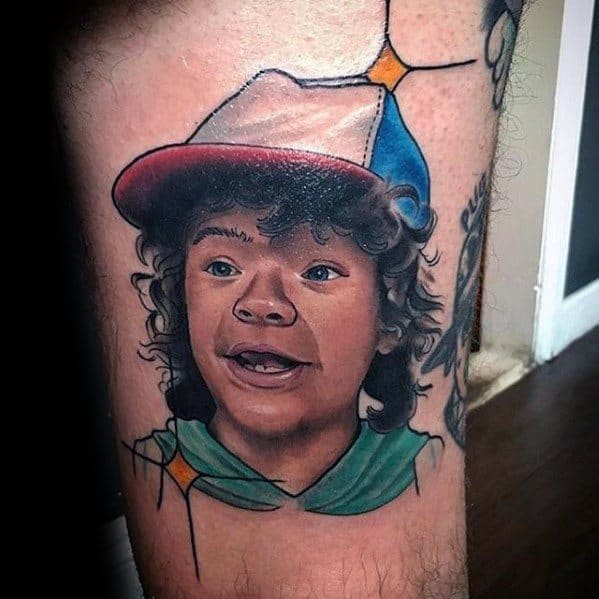 Realistic color tattoo of Dustin from Stranger Things with a surprised expression, wearing his iconic cap and green hoodie.