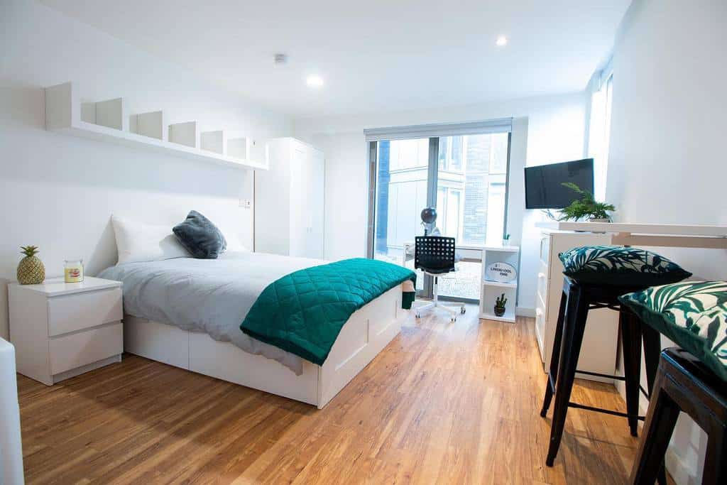 Spacious studio apartment bedroom with white furniture, desk area, and wooden floors.