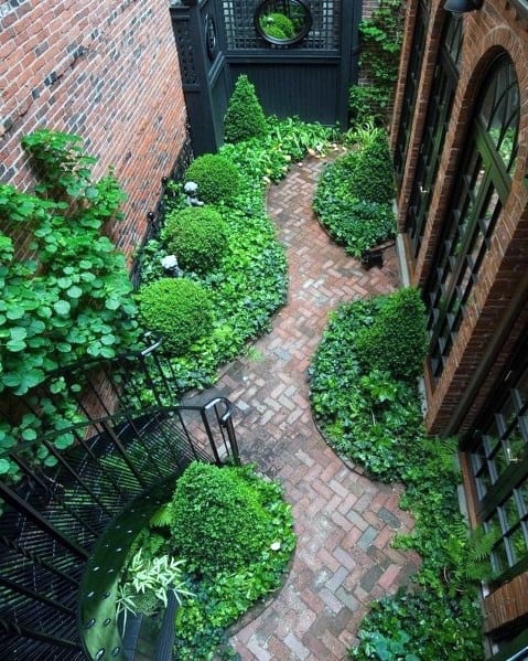 A narrow brick walkway winds through a lush, green garden beside a brick building