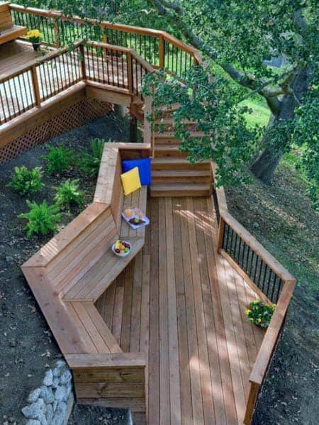 Wooden deck with built-in bench seating, stairs, and potted plants under a large tree