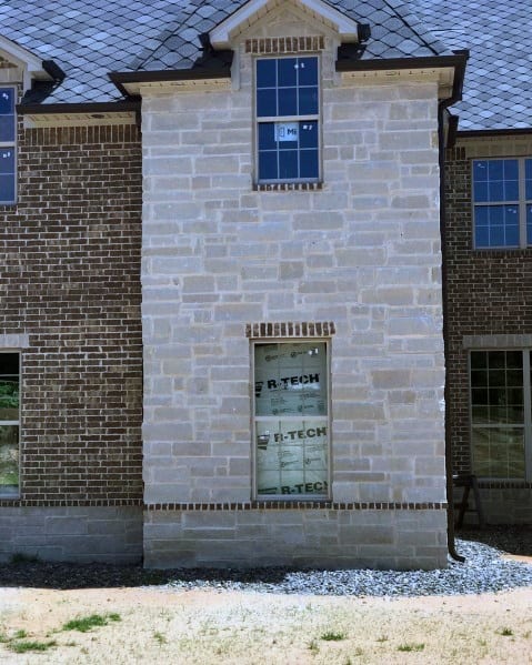 Modern home under construction featuring a mix of light stone and brick cladding, black-framed windows, and a steep gabled roofline