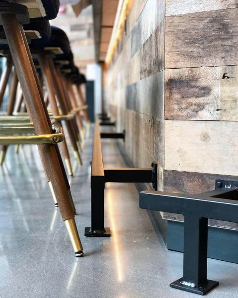 Square metal tube foot rail attached to a rustic wooden bar with brass-accented stools on polished concrete flooring.