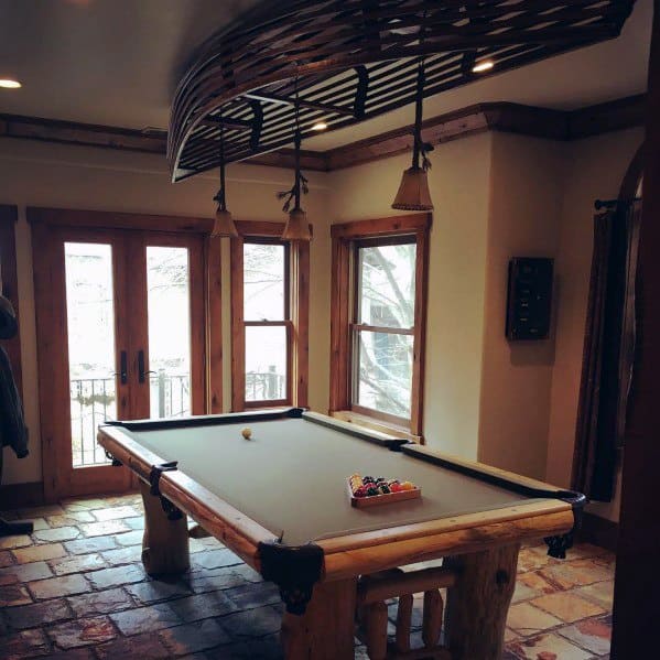 Rustic billiard room with log-style pool table, stone flooring, and natural wood-framed windows.