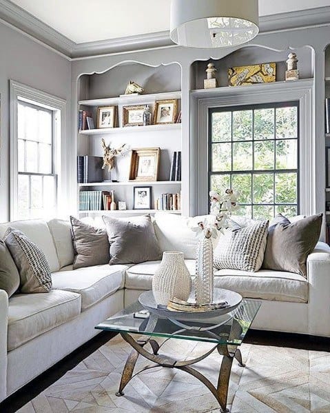 Cozy living room with built-in bookcases framing large windows, featuring a white sectional sofa, soft grey walls, decorative pillows, and a glass-top coffee table