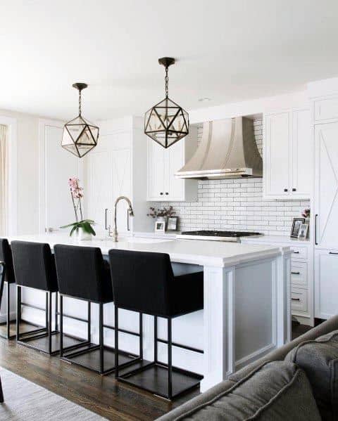 White kitchen with black bar stools, geometric pendant lights, and subway tile backsplash.