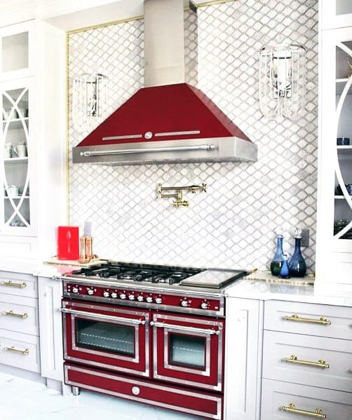 Elegant kitchen with a red range and hood, diamond-patterned stone backsplash, and gold-accented white cabinetry