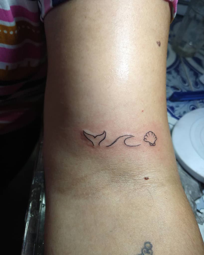 Minimalist ocean-inspired tattoo featuring a whale tail, a wave, and a small seashell in fine line design