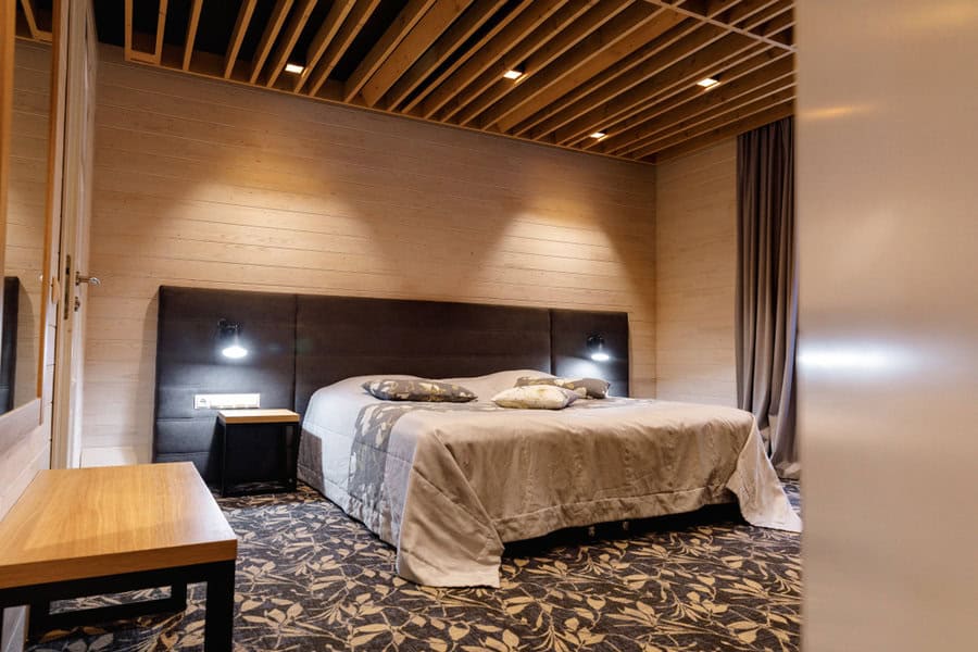 A modern bedroom with a large bed, soft lighting, wooden ceiling beams, and patterned carpeting, plus a small wooden table and nightstand beside the bed