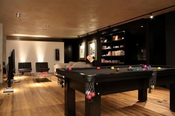 A stylish game room with a pool table, cozy seating, bookshelves, and wooden floors, softly lit for ambiance