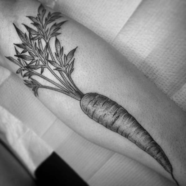 Realistic black and gray carrot tattoo on the forearm with fine line detailing, intricate shading, and lush leafy tops for a bold botanical look.