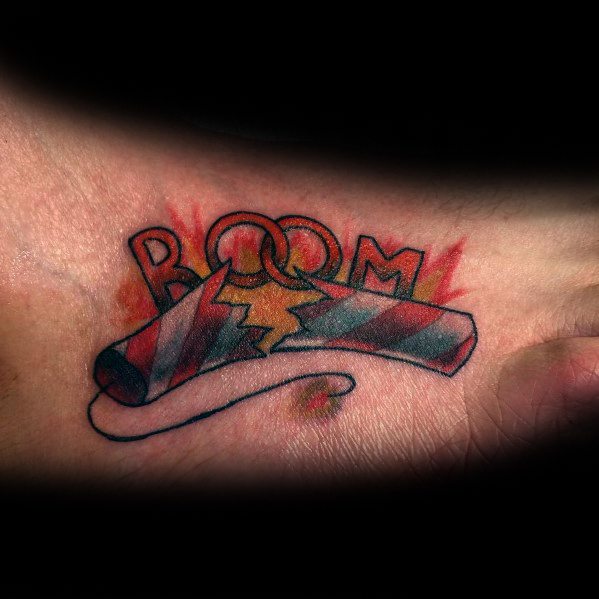 Tattoo of a broken firecracker with a bold "BOOM" text behind it, featuring red and blue shading and explosive details for a dynamic design.