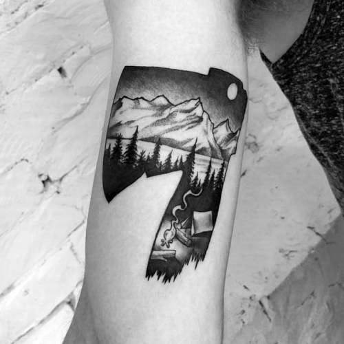 Black and grey hatchet tattoo with a detailed landscape scene inside the blade, featuring mountains, pine trees, a campfire, and a moonlit sky.