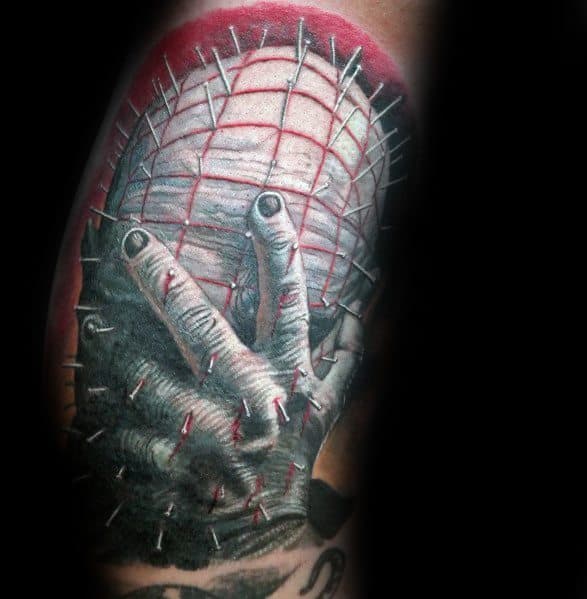 Realistic *Hellraiser* tattoo of Pinhead with a hand gripping his face, featuring deep shading, red accents, and intricate pin details for a haunting effect.