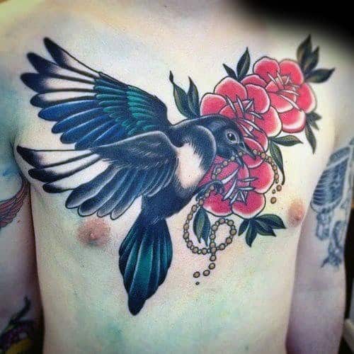 Colorful magpie chest tattoo featuring the bird in flight with vibrant blue feathers, holding a pearl necklace near red flowers for a bold design.