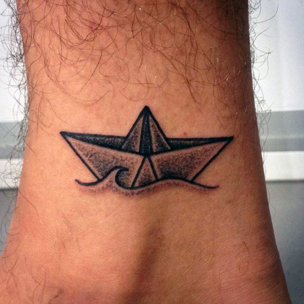 Black ink tattoo of a paper boat floating on waves, placed on the ankle, symbolizing adventure, resilience, and life’s journey.