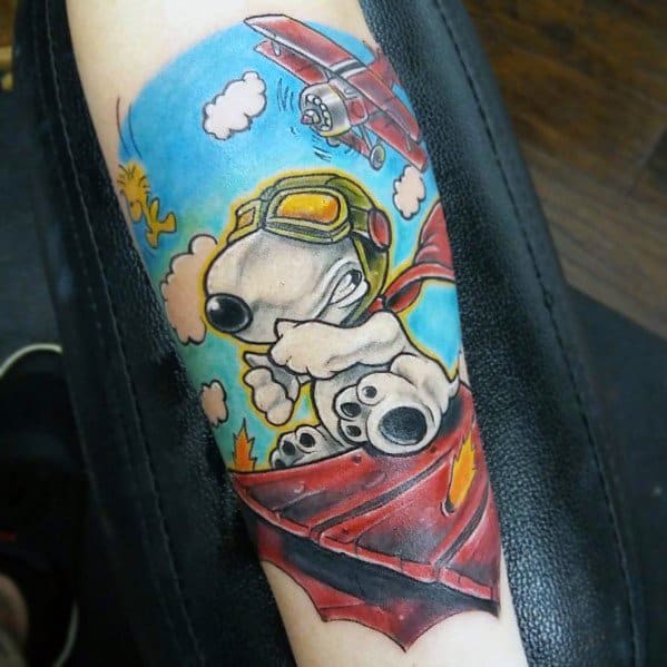 Colorful tattoo of Snoopy as a flying ace, wearing goggles and a red scarf, battling a plane in the sky while atop his burning red doghouse.