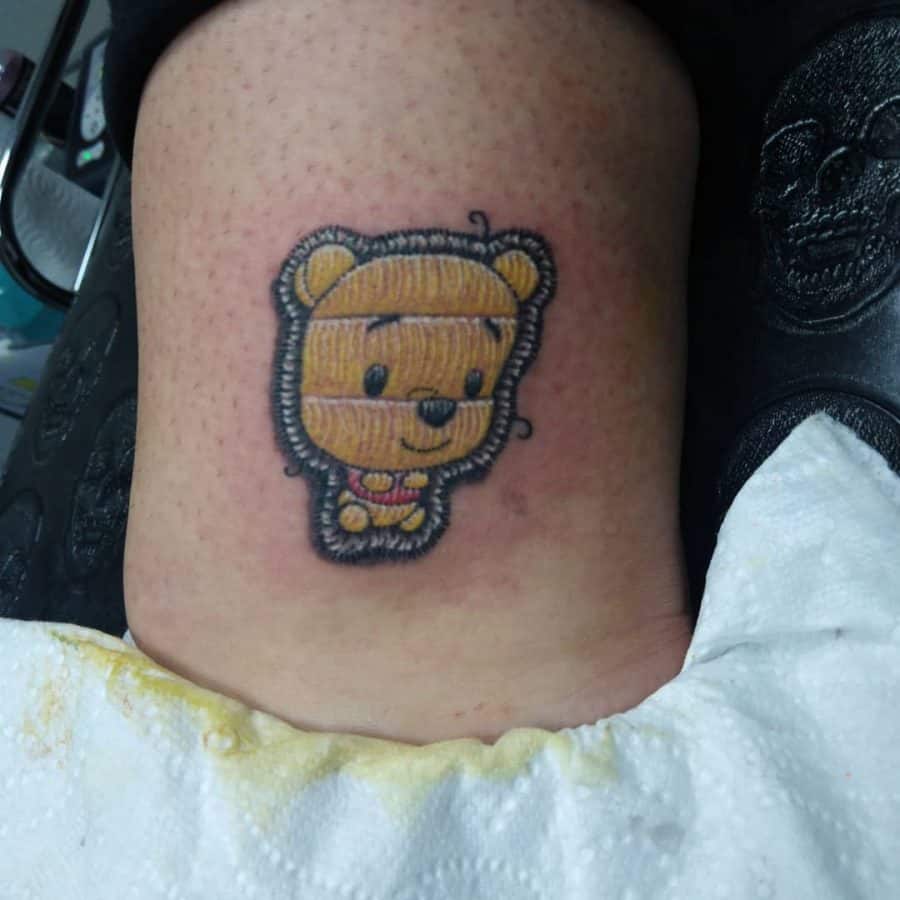 Tattoo of a small, cute Pooh Bear with an embroidered quilted texture, giving it the look of a sewn-on patch.