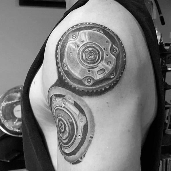 Realistic black and grey Subaru-themed tattoo on the upper arm, featuring detailed flywheel and clutch components with shading for a 3D effect.