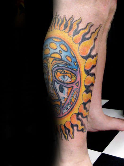 Vibrant Sublime sun face tattoo wrapping around the calf, with bold orange and yellow flames and detailed blue-toned facial features.