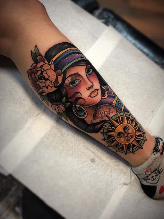 Vibrant tattoo of a gypsy woman with a floral headpiece, detailed jewelry, and a sun and moon design on the lower leg.