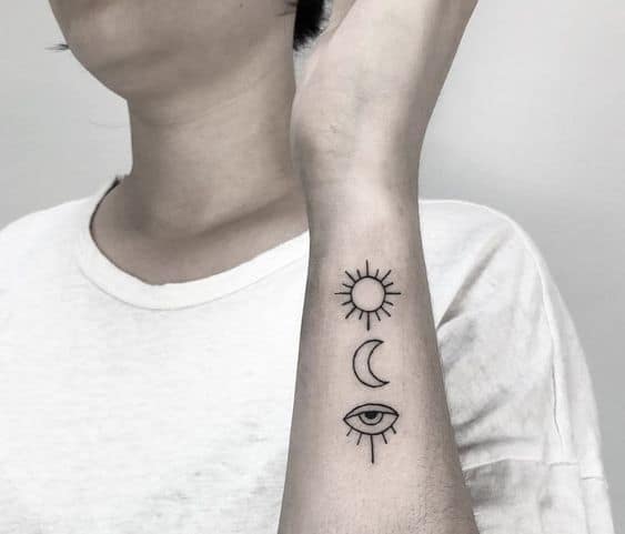 Minimalist tattoo of a sun, crescent moon, and all-seeing eye, inked in fine black lines on the forearm.