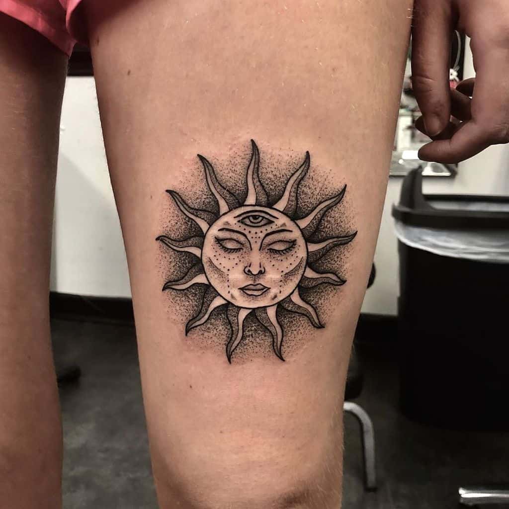 Tattoo of a sun with a serene face and a third eye, surrounded by radiant flames, inked in black and dot shading on the thigh.