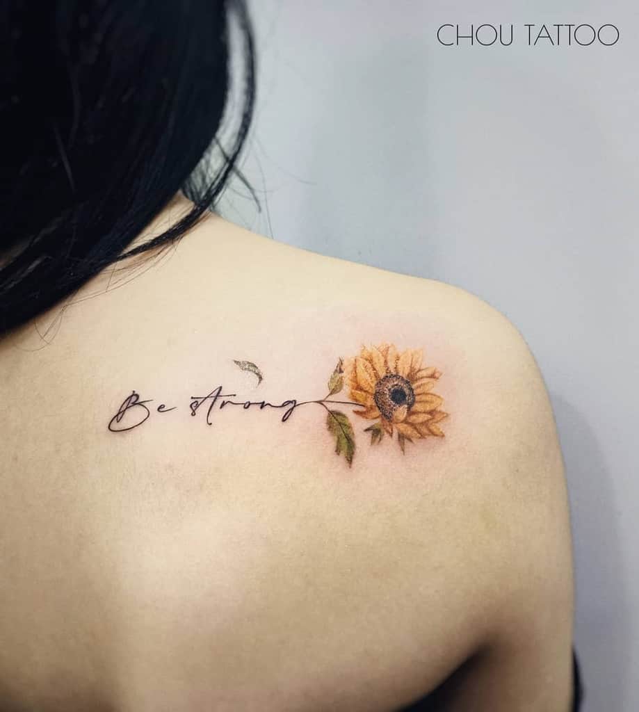 A tattoo with "Be Strong" elegantly written in cursive and a vibrant sunflower graces the upper shoulder