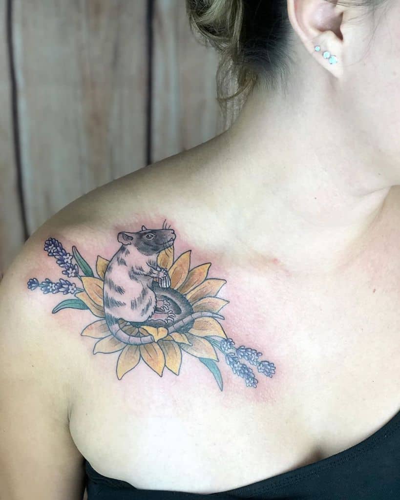 A tattoo on the shoulder featuring a small rat sitting amidst a vibrant sunflower with lavender sprigs, blending realistic and illustrative styles.