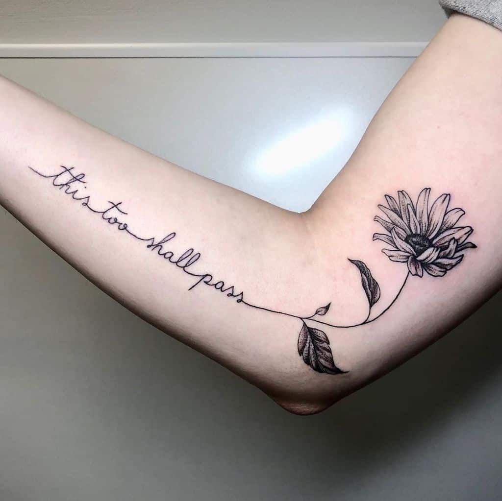 A flowing "This too shall pass" tattoo in cursive along the inner arm, seamlessly turning into a delicate daisy, symbolizing growth and perseverance.