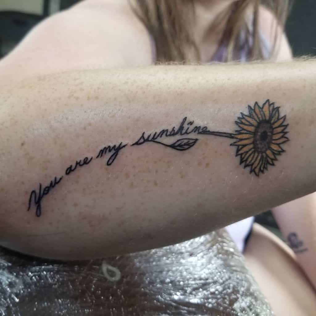 Minimalist tattoo featuring 'You are my sunshine' in script with a delicate sunflower and leaf accent on the arm