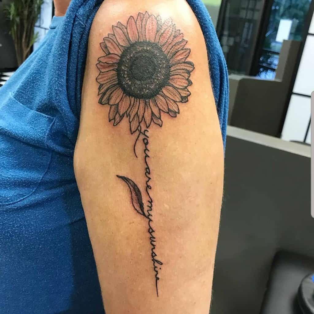 Sunflower tattoo on upper arm with "you are my sunshine" text as the stem