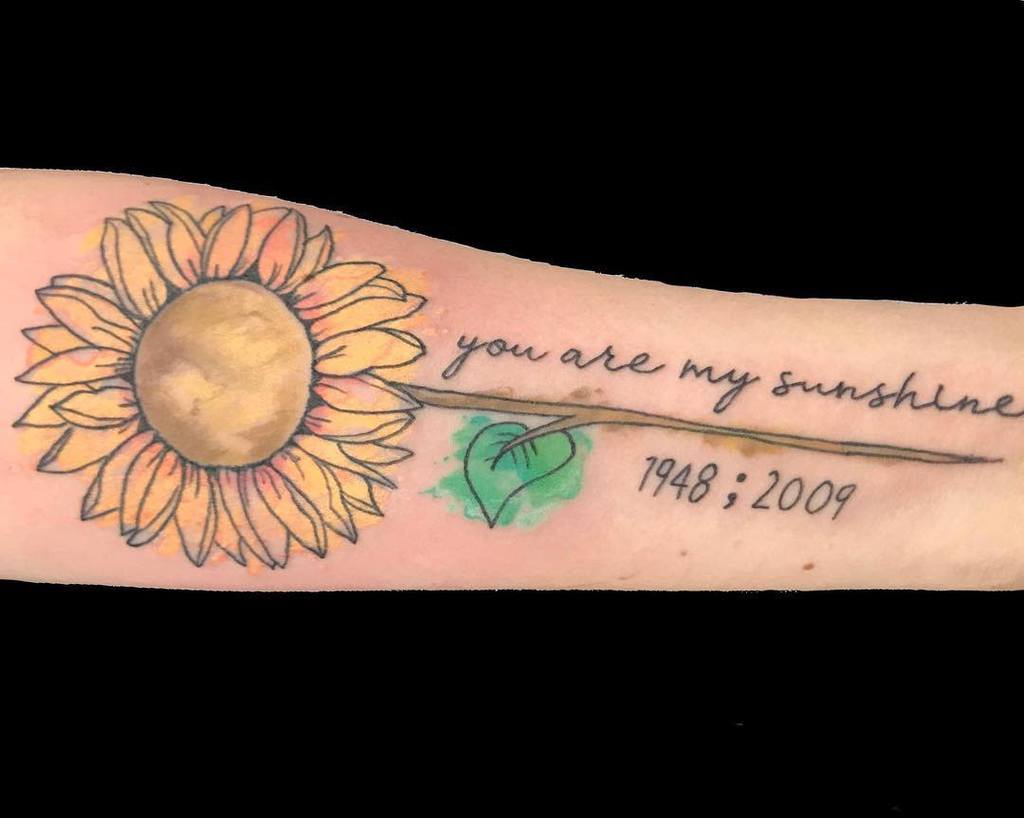 A sunflower tattoo on an arm with text: "you are my sunshine" and dates 1948; 2009