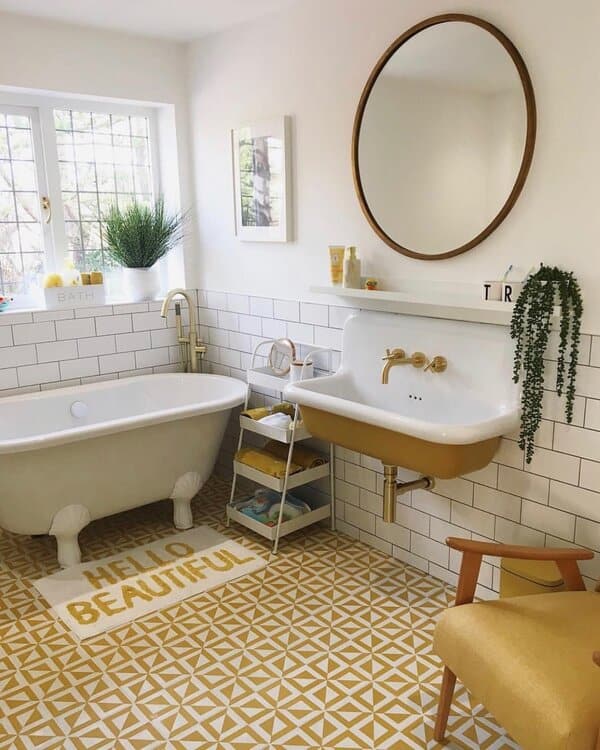 White bathroom with mustard sink, gold fixtures, geometric floor tiles, and a clawfoot tub under a round wood-framed mirror.