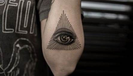 Tattoo of an all-seeing eye inside a triangle with radiating lines, inked in black on the forearm.