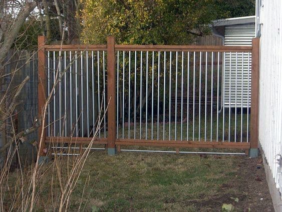 A fenced metal gate with wooden posts in a grassy backyard, surrounded by trees and a white building