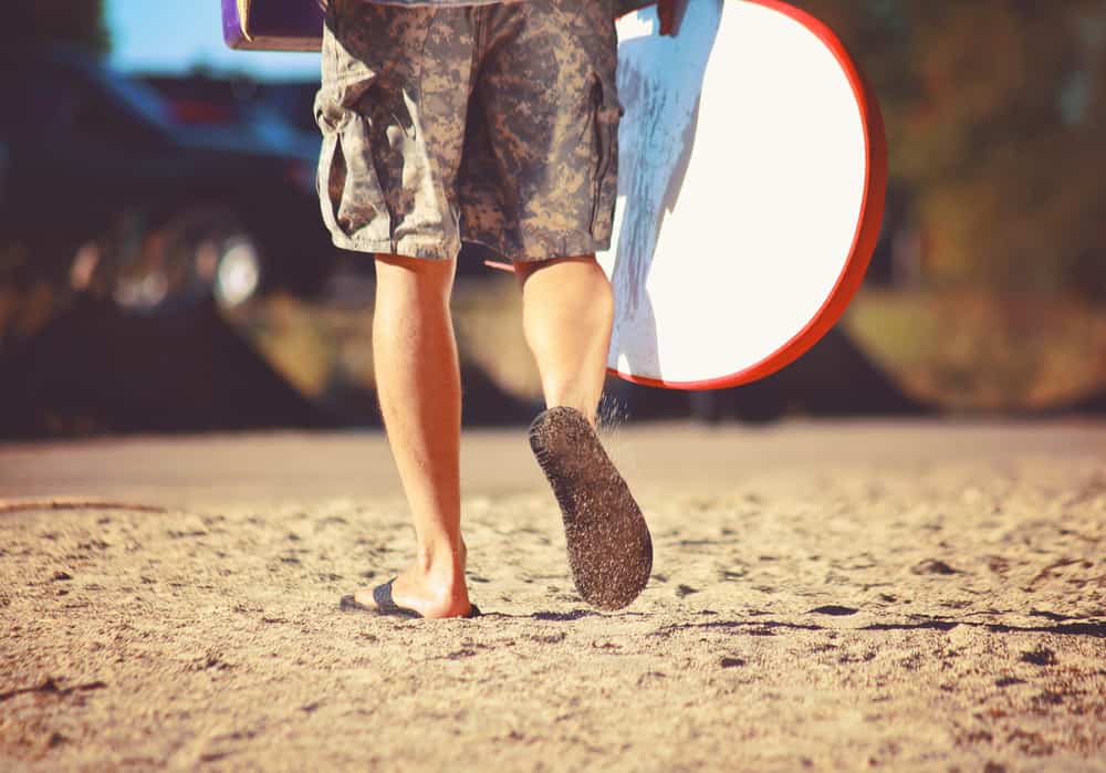A surfer walking on the beach wearing flip-flops and carrying a surfboard