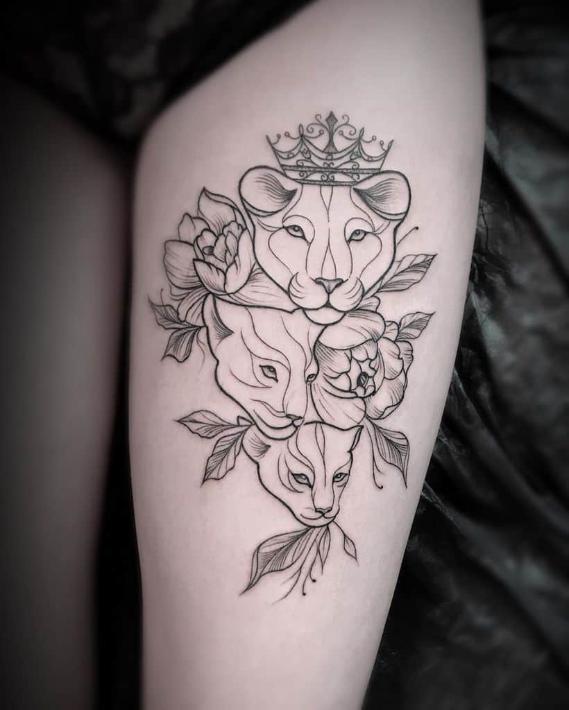 Elegant black linework tattoo on the thigh, featuring three lioness faces, floral accents, and a crown, symbolizing strength and femininity.