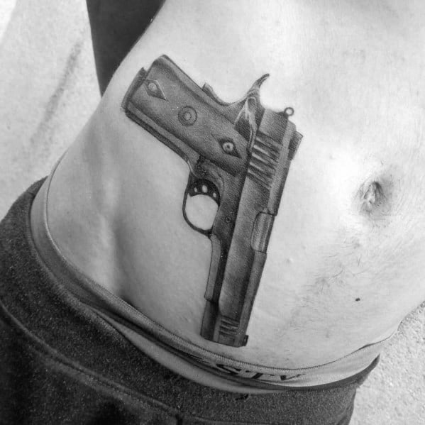 Realistic black and gray tattoo of a 1911 pistol on the lower abdomen, designed to appear as if tucked into the waistband for a three-dimensional effect.