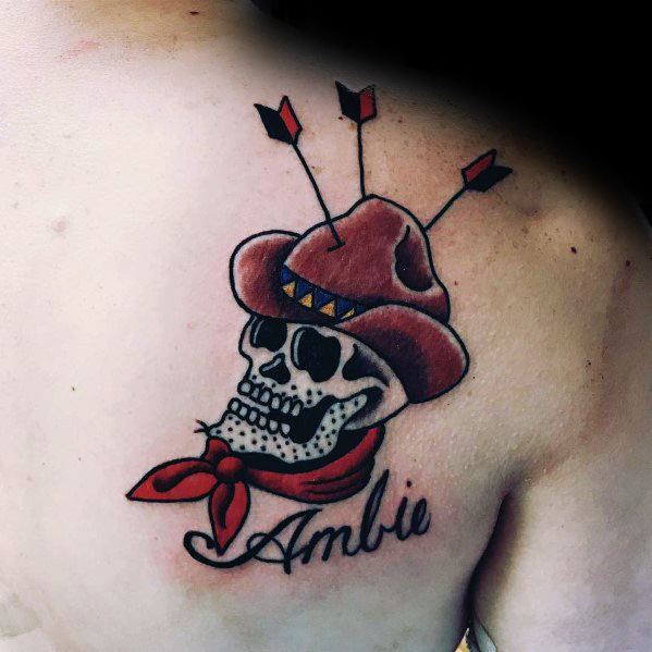 Tattoo of a cowboy skull wearing a red hat and bandana, pierced by three arrows, with the name "Ambie" written below in cursive script.