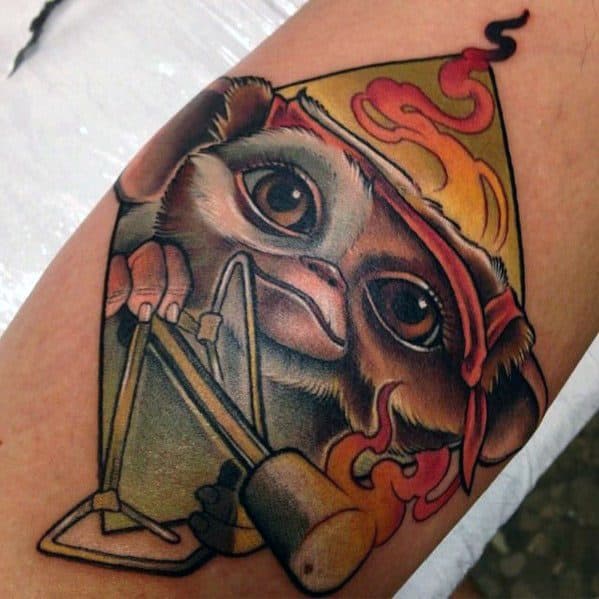 Vibrant Gizmo tattoo holding a slingshot, wearing a flaming party hat, blending cuteness with a mischievous, playful twist.