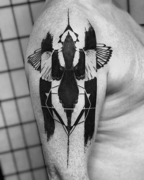 Geometric magpie tattoo on upper arm, featuring symmetrical patterns, bold black brush strokes, and intricate wing details for a modern look.