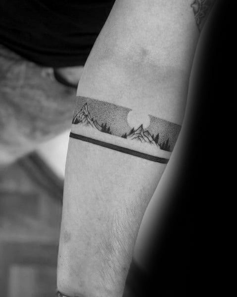 Minimalist mountain armband tattoo on the forearm, featuring fine dot shading, pine trees, and a sun or moon for a sleek and nature-inspired design.