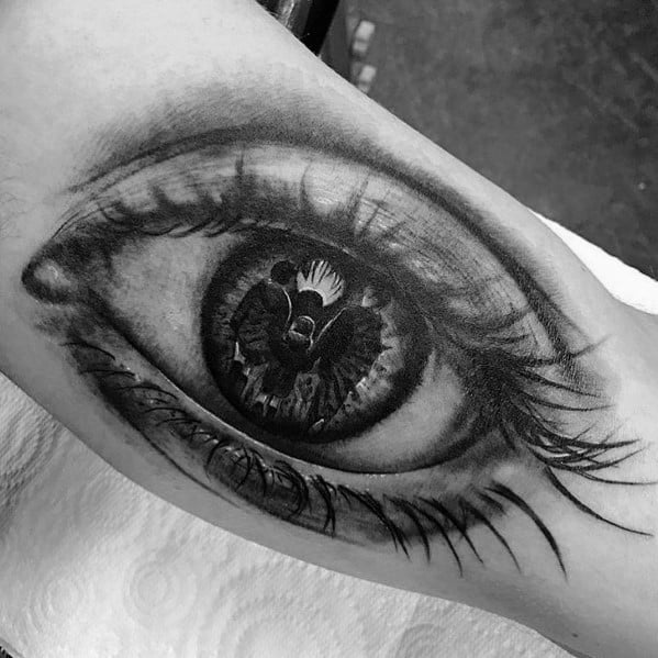 Tattoo of a realistic eye with a reflection of an angel and a kneeling figure, creating a deep and symbolic design with intricate shading.
