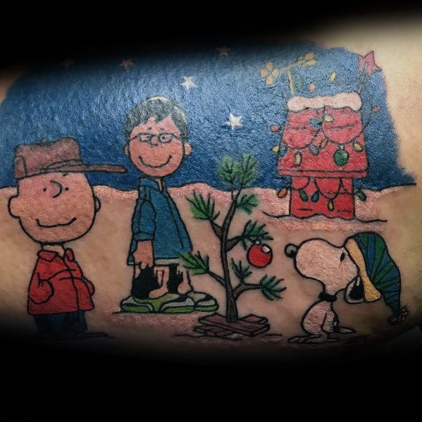 Colorful tattoo of Charlie Brown, Linus, and Snoopy gathered around a small Christmas tree, with Snoopy’s decorated doghouse in the background.