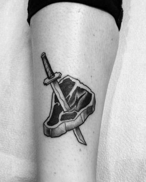 Black and gray tattoo of a steak pierced by a dagger, featuring bold shading and fine details for a classic and striking design.