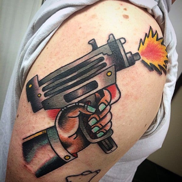 Bold neo-traditional Uzi tattoo on the upper arm, featuring a hand gripping the gun with vibrant colors, thick outlines, and a firing effect.