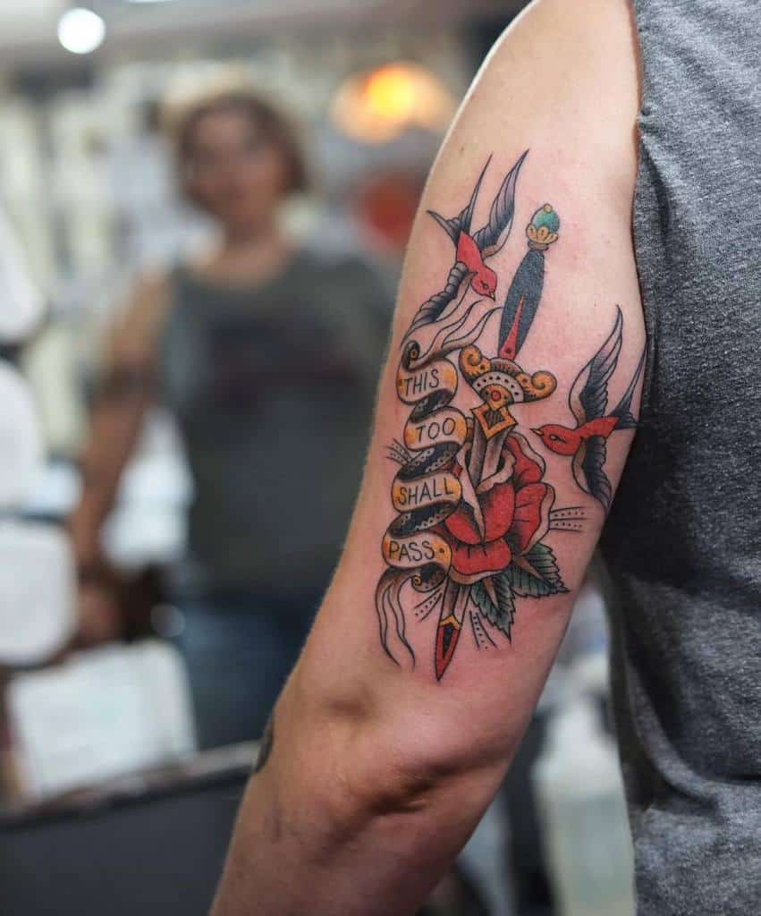 This tattoo design features the phrase "This too shall pass" surrounded by vibrant imagery, including two birds and a blooming rose, with a decorative banner and colorful elements. The design combines traditional tattoo art with an inspirational message, creating a bold, striking look.
