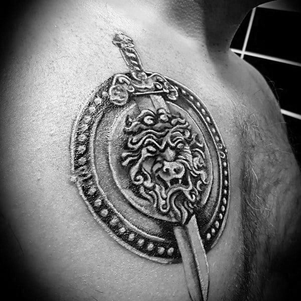 Chest tattoo of a detailed lion head on a circular shield with a beaded border, pierced by a vertically positioned ornate sword.