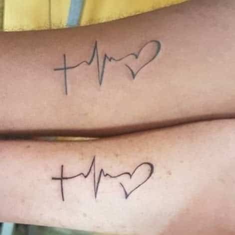 Two arms with matching tattoos depicting a cross, heartbeat line, and heart design