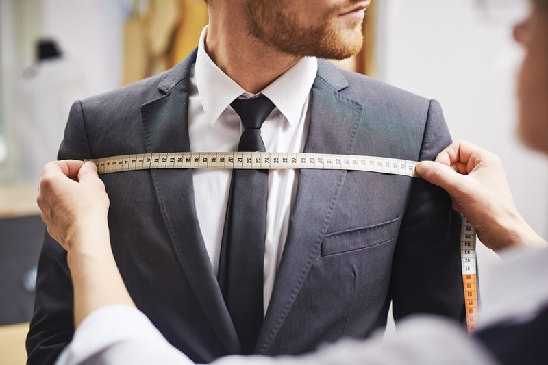 A tailor measures the chest of a man wearing a dark grey suit and black tie